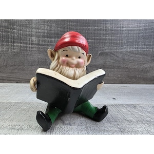 Puede incluir: Una figura de gnomo de jardín de cerámica leyendo un libro. El gnomo lleva un gorro rojo, pantalones verdes y zapatos negros. El libro tiene una cubierta negra y páginas blancas. El gnomo tiene barba blanca y mejillas rosadas.