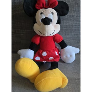May include: A plush Minnie Mouse doll. The doll has a red dress with white polka dots, a red bow, and yellow shoes. The doll's face is white with black eyes, a black nose, and a smiling expression. The doll is sitting.