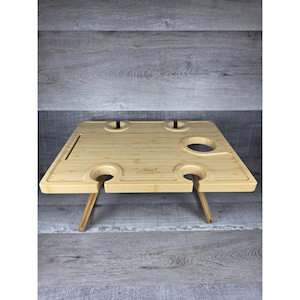 May include: A light-colored bamboo picnic table with two wine glass holders and two bottle holders. The table has two folding legs and a small groove for a phone or other item. The table is 15 inches wide and 12 inches long.