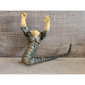Könnte beinhalten: Detaillierte Actionfigur eines Monsters mit reptilienartigem Körper und erhobenen Armen. Die Figur hat einen beigefarbenen Kopf mit einem bedrohlichen Grinsen, der in einen dunkelblauen und grauen Körper übergeht. Der Schwanz ist lang und segmentiert.