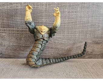 May include: A detailed action figure of a monster with a reptilian body and raised arms. The figure has a tan head with a menacing grin, transitioning to a dark blue and gray body. The tail is long and segmented.