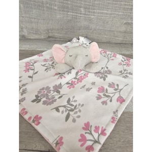 May include: A white baby blanket with a pink and grey floral pattern. A grey elephant with pink ears is attached to the blanket.