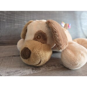 Puede incluir: Un juguete de peluche con forma de perro en tonos beige y marr&oacute;n. El perro tiene orejas ca&iacute;das, una nariz marr&oacute;n y una mancha marr&oacute;n sobre un ojo. Est&aacute; tumbado sobre una superficie de madera. Un peque&ntilde;o lazo colorido est&aacute; atado a la oreja del perro.