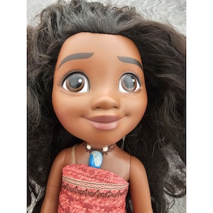Puede incluir: Una mu&ntilde;eca de piel morena con cabello negro largo que lleva un vestido rojo y marr&oacute;n y un collar azul con un colgante de concha.