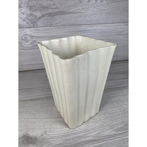 May include: A cream-colored ceramic vase with a square opening and a tapered base. The exterior features a series of vertical, rounded fluted ridges that extend from the top rim down to the bottom, creating a textured, scalloped appearance.