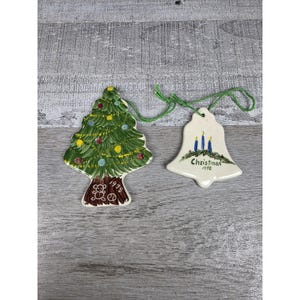 May include: Two ceramic ornaments with green string hangers. One is a green Christmas tree with yellow and red ornaments and a brown trunk marked 1978. The other is a white bell featuring three blue candles and the text Christmas 1998.