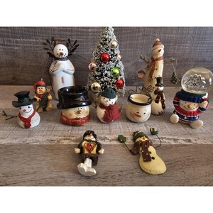 May include: A collection of festive snowman figurines and Christmas decorations. The assortment includes snowmen with top hats, scarves, and a snow globe. A small Christmas tree with ornaments is also present.