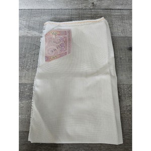 May include: A white fabric square with a pink and white label that says "K.Kee". The fabric has a slightly textured surface and is surrounded by a light brown border.