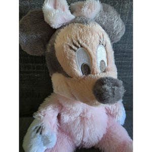 May include: A plush Minnie Mouse doll with a pink and gray color scheme. The doll features large, embroidered eyes, a gray nose, and soft, fluffy fur. The doll's ears are gray with pink accents, and the body is pink.