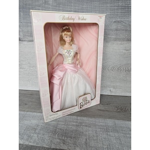 Może przedstawiać: Lalka Barbie w białej i różowej sukience z kwiatowym wzorem. Lalka znajduje się w pudełku z napisem "Birthday Wishes" i "Barbie". Pudełko jest różowe i białe w kratkę.