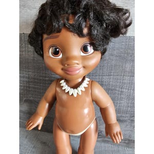 Pu&ograve; includere: Una bambola di pelle marrone con capelli neri ricci, che indossa una collana di conchiglie bianche e un fondo bikini marrone chiaro. La bambola ha gli occhi marroni e un'espressione sorridente.