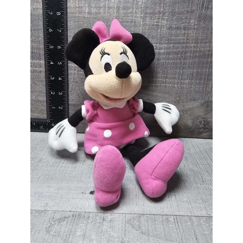 May include: A plush Minnie Mouse doll, featuring black ears, a pink bow, and a pink dress with white polka dots. The doll has white gloves and pink shoes. The doll is sitting down.