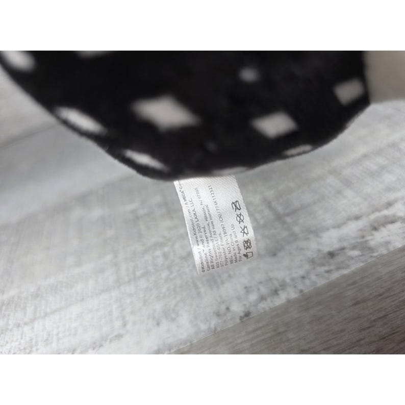 May include: A close-up of a black and white plush toy with a tag. The plush toy has a pattern of white diamonds on a black background. The tag includes text about the manufacturer and care instructions.