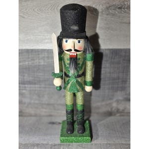 May include: A decorative nutcracker soldier figurine. The figurine is dressed in a green and gold uniform with a black top hat and holds a wooden sword. The base is green and glittery.