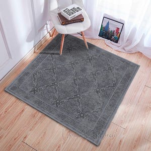 Può includere: Un tappeto quadrato grigio con un motivo floreale ornato. Il tappeto è posizionato su un pavimento in legno, con una sedia bianca e un'immagine incorniciata sullo sfondo. Il tappeto ha un bordo e un design dettagliato.