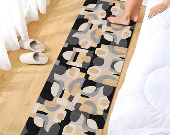 May include: A rectangular rug with an abstract pattern in black, gray, and beige tones. The rug is placed on a wooden floor, with a bed and slippers visible in the background. The design features geometric shapes.