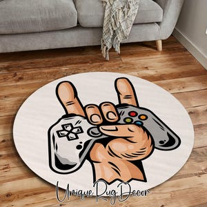 Pode incluir: Um tapete redondo com uma ilustração de desenho animado de uma mão segurando dois controles de videogame. A mão faz o sinal de rock and roll. O tapete é branco sujo com o texto "Unique Rug Decor" em fonte cursiva.