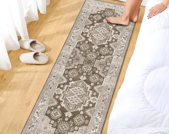 May include: A rectangular, patterned rug in shades of brown, beige, and cream. The rug features a central geometric design with intricate borders. It is placed on a wooden floor, near a bed with white bedding and a pair of slippers.
