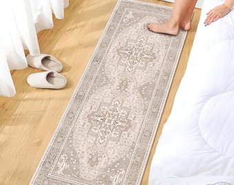 May include: A rectangular, beige and gray patterned runner rug with an ornate, symmetrical design. The rug is placed on a wooden floor next to a bed with white bedding. The rug's dimensions are approximately 2.5 feet wide and 6 feet long.