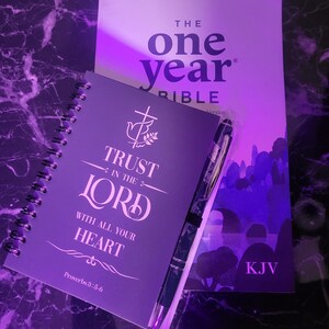 May include: A purple journal with the words "Trust in the Lord with all your heart" and "Proverbs 3:5-6" printed on the cover. The journal is next to a purple Bible with the words "The One Year Bible" and "KJV" printed on the cover.