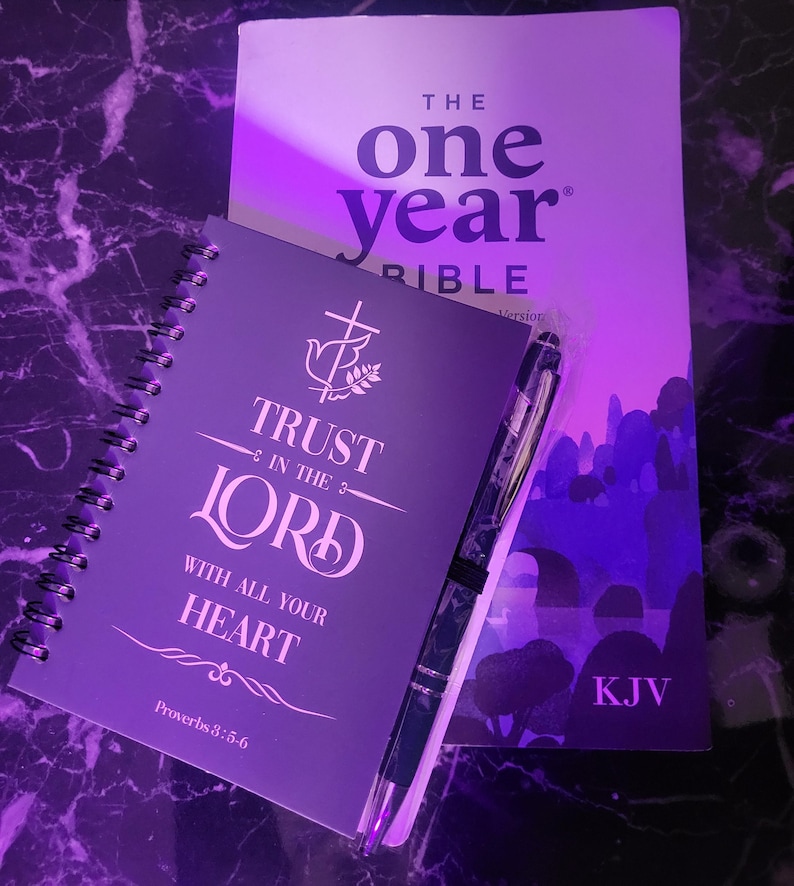 May include: A purple journal with the words "Trust in the Lord with all your heart" and "Proverbs 3:5-6" printed on the cover. The journal is next to a purple Bible with the words "The One Year Bible" and "KJV" printed on the cover.