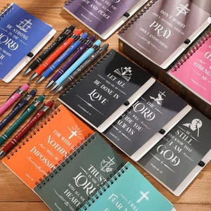 May include: A collection of small, spiral bound notebooks with inspirational quotes and bible verses. The notebooks are in various colors, including black, gray, orange, green, blue, and pink. Each notebook features a cross design and a different quote. The quotes include "Be strong in the Lord", "Let all that you do be done in love", "The Lord will guide you always", "For with God nothing shall be impossible", "Trust in the Lord with all your heart", "Be still and know that I am God", "Don't worry about anything instead pray about everything", and "Strength in me".