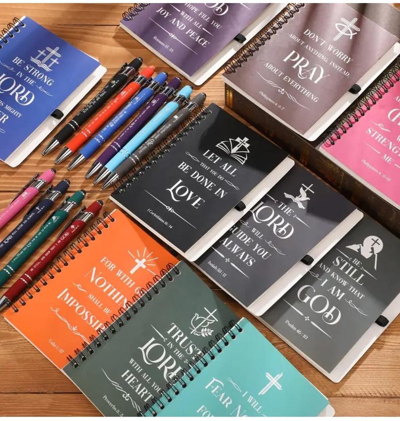 May include: A collection of small, spiral bound notebooks with inspirational quotes and bible verses. The notebooks are in various colors, including black, gray, orange, green, blue, and pink. Each notebook features a cross design and a different quote. The quotes include "Be strong in the Lord", "Let all that you do be done in love", "The Lord will guide you always", "For with God nothing shall be impossible", "Trust in the Lord with all your heart", "Be still and know that I am God", "Don't worry about anything instead pray about everything", and "Strength in me".