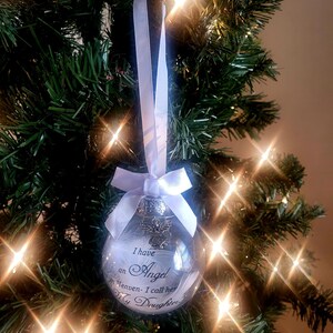 May include: White glass Christmas ornament with a white ribbon and a silver angel charm. The ornament has the text "I have an Angel in Heaven - I call her My Daughter".