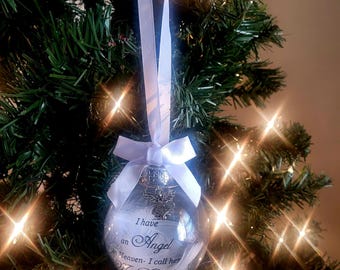 Op de afbeelding: Witte glazen kerstbal met een wit lint en een zilveren engelbedeltje. De bal heeft de tekst "Ik heb een engel in de hemel - Ik noem haar mijn dochter".