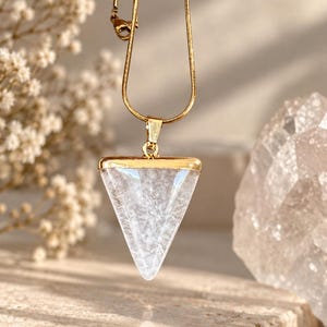 May include: A gold-toned chain necklace featuring a triangular pendant with a clear, textured stone insert. The pendant has a smooth gold-coloured metal top edge and is suspended from a thin, flexible gold-toned snake chain with a metal clasp.