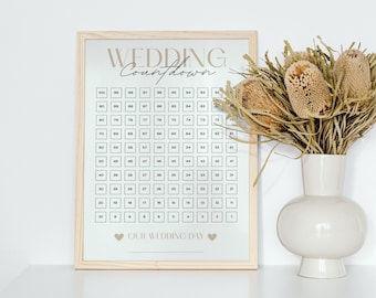 May include: A wooden framed print with a white background and gold text that says "Wedding Countdown". The print features a grid of numbers from 100 to 1, with the text "Our Wedding Day" at the bottom.