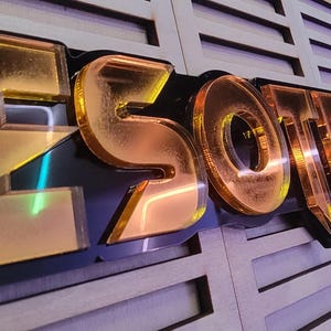 May include: A gold-colored, three-dimensional sign that reads "ESOTERIC" in bold, stylized letters. The letters are mounted on a black base, with a wooden panel background. The sign has a reflective, metallic finish.