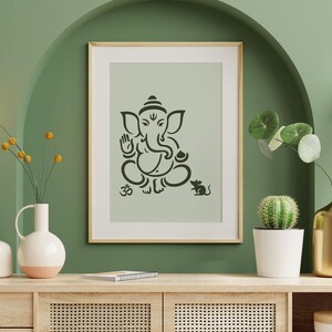May include: A framed print featuring a black silhouette of the Hindu deity Ganesha, with the Om symbol and a mouse. The artwork is displayed on a light green background, with a wooden frame. The room is decorated with plants and vases.