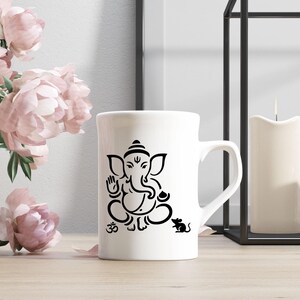 May include: White ceramic mug featuring a black illustration of the Hindu deity Ganesha, with the Om symbol and a mouse. The mug has a curved handle and is set on a wooden surface, with pink flowers in the background.