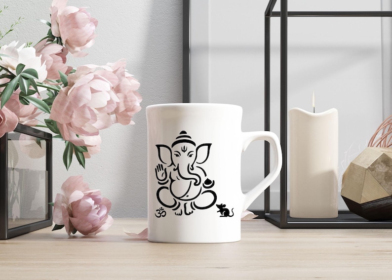 May include: White ceramic mug featuring a black illustration of the Hindu deity Ganesha, with the Om symbol and a mouse. The mug has a curved handle and is set on a wooden surface, with pink flowers in the background.