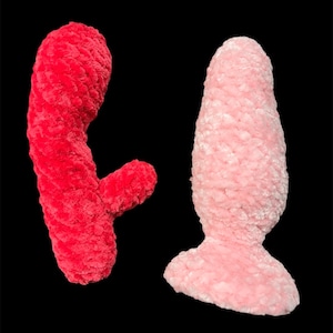 May include: Two plush sex toys. One is a vibrant red, curved shape with a smaller appendage. The other is a soft pink, tapered shape with a flared base. Both are made of a textured, fuzzy material against a black background.
