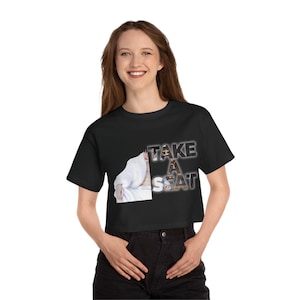 May include: Black crop top with the text "TAKE A SEAT" in a stylized font. The shirt features a graphic of a person in a white robe. The shirt is made of a soft material.