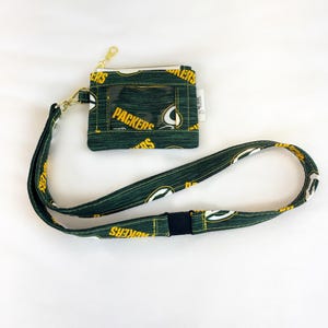 Pode incluir: Carteira de tecido verde e amarelo com uma janela de identificação transparente e um zíper dourado, presa a um cordão correspondente. A carteira e o cordão apresentam um padrão repetido da palavra "Packers" e um logotipo.