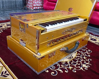May include: A gold-coloured harmonium with a keyboard and bellows. The harmonium is open and the keyboard is visible. The harmonium is sitting on a red rug with a patterned design.