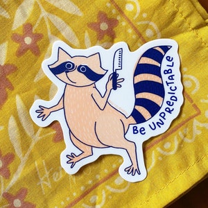 May include: A white sticker with a cartoon raccoon holding a knife. The raccoon has a pink body, black eyes, and a blue and white striped tail. The text "Be Unpredictable" is written vertically on the right side of the sticker.