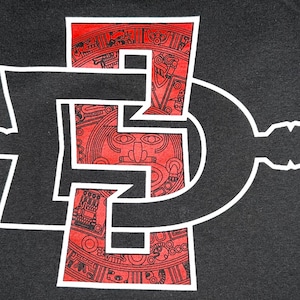 Peut inclure: Un design graphique noir et blanc d'une lettre stylisée "D" avec un motif aztèque rouge et blanc à l'intérieur. Le design est sur un fond noir.