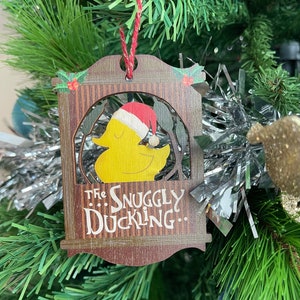 May include: A wooden Christmas bauble shaped like a house with a yellow duck wearing a red Santa hat inside. The bauble has the text "...The Snuggly Duckling..." on the bottom.