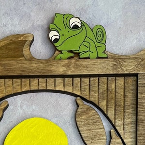 May include: A wooden wall decoration featuring a green chameleon with large eyes, perched on a wooden ledge. The chameleon is looking to the left, and the ledge is part of a larger wooden structure with a yellow circle and a dark outline.