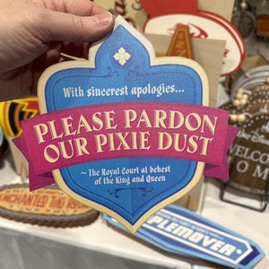 May include: A shield-shaped sign with a blue background and a pink banner across the centre. The text reads: With sincerest apologies... PLEASE PARDON OUR PIXIE DUST ~ The Royal Court at behest of the King and Queen. The sign features decorative scrollwork.