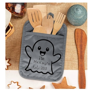 May include: A gray pot holder with a ghost design and the text "The McKay's Est. 2017". The pot holder contains wooden kitchen utensils. A wooden spoon and a decorative blue ball are also visible.