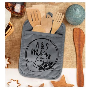 May include: A gray pot holder with a pocket holding wooden kitchen utensils. The pot holder features a black graphic of a cowboy hat, a flower, and the text "A & S McKay" with the date "05.23.17" inside a circle.