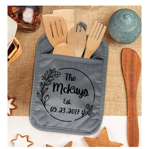 May include: A gray pot holder with a pocket, holding wooden kitchen utensils. The pot holder has a black floral design with the text "The McKays Est. 05.23.2017" inside a circle. Other kitchen items are in the background.