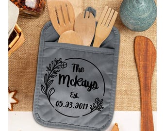 May include: A gray pot holder with a pocket, holding wooden kitchen utensils. The pot holder has a black floral design with the text "The McKays Est. 05.23.2017" inside a circle. Other kitchen items are in the background.