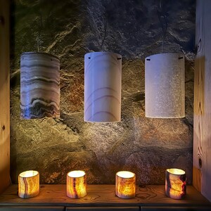 Op de afbeelding: Drie hangende lichtarmaturen gemaakt van wit en bruin albast, met een warme gloed. Daaronder bevinden zich vier kleinere albast kandelaars, ook aangestoken, op een houten plank.