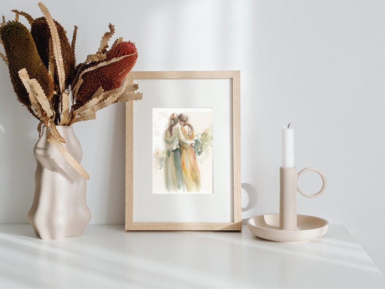 May include: A framed watercolor painting of two women embracing, with a soft, muted color palette of greens, yellows, and browns. The painting is set against a white background and is displayed on a white shelf with a candle and vase of dried flowers.